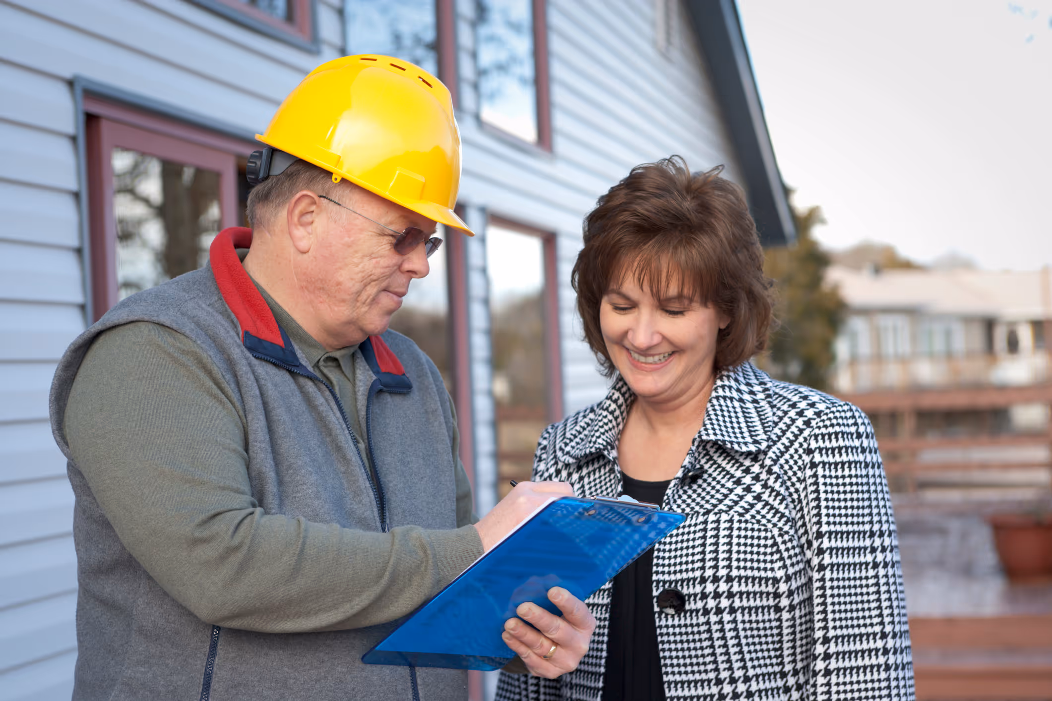 A contractor in a hard hat reviews a written estimate on a clipboard with a homeowner outside a residential property.